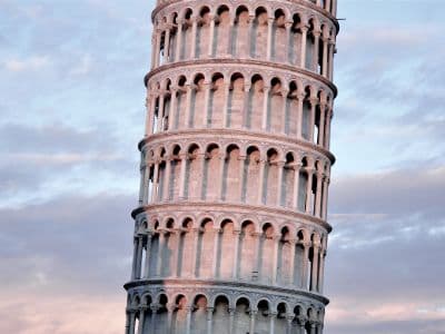 A detailed view of the Leaning Tower of Pisa, capturing its significant tilt. The cylindrical structure is visible, with several levels featuring intricate open arcades supported by columns. The light-colored stone contrasts with a sky filled with soft clouds.