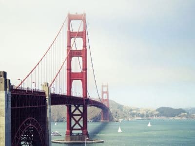 A partial view of the Golden Gate Bridge's southern tower and cables is shown, with the full bridge extending into the distance across the water. The famous international orange suspension bridge stands against a hazy sky, with sailboats and other vessels on the water below and green hills in the background.